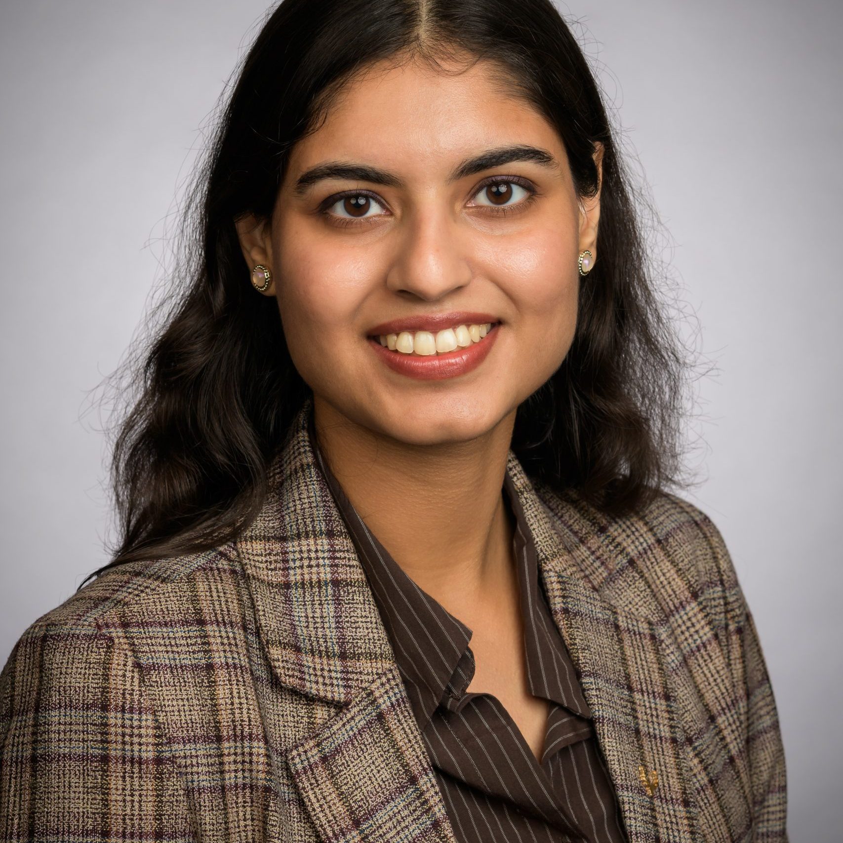 Portrait of a smiling woman with dark hair, wearing a brown striped blouse and plaid blazer, against a neutral gray backdrop.