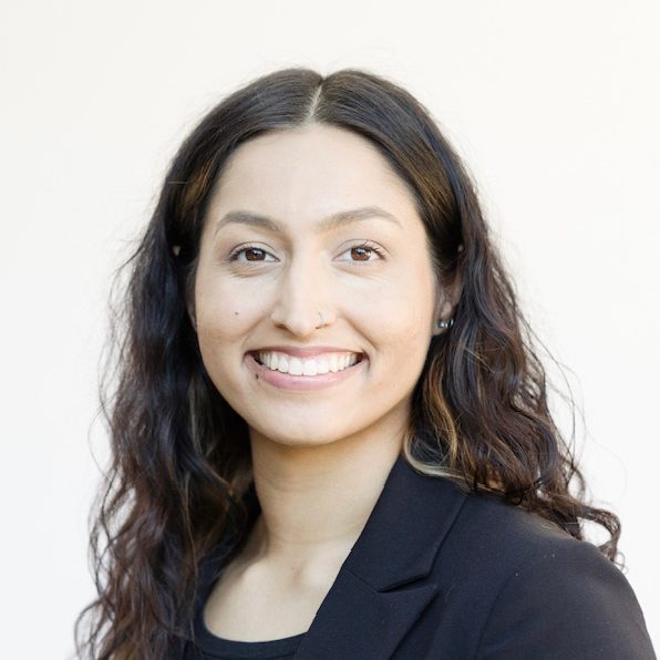 Professional headshot of a smiling woman with long dark wavy hair wearing a black blazer against a light background.